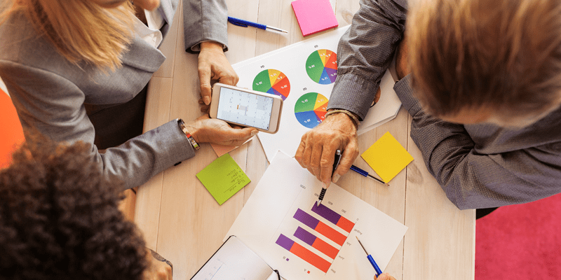 A photo of three people reviewing some data on the pieces of paper on the table in front of them. There are colorful bar charts and pie charts on the table. One person is holding a phone, and the other two are holding pens. 