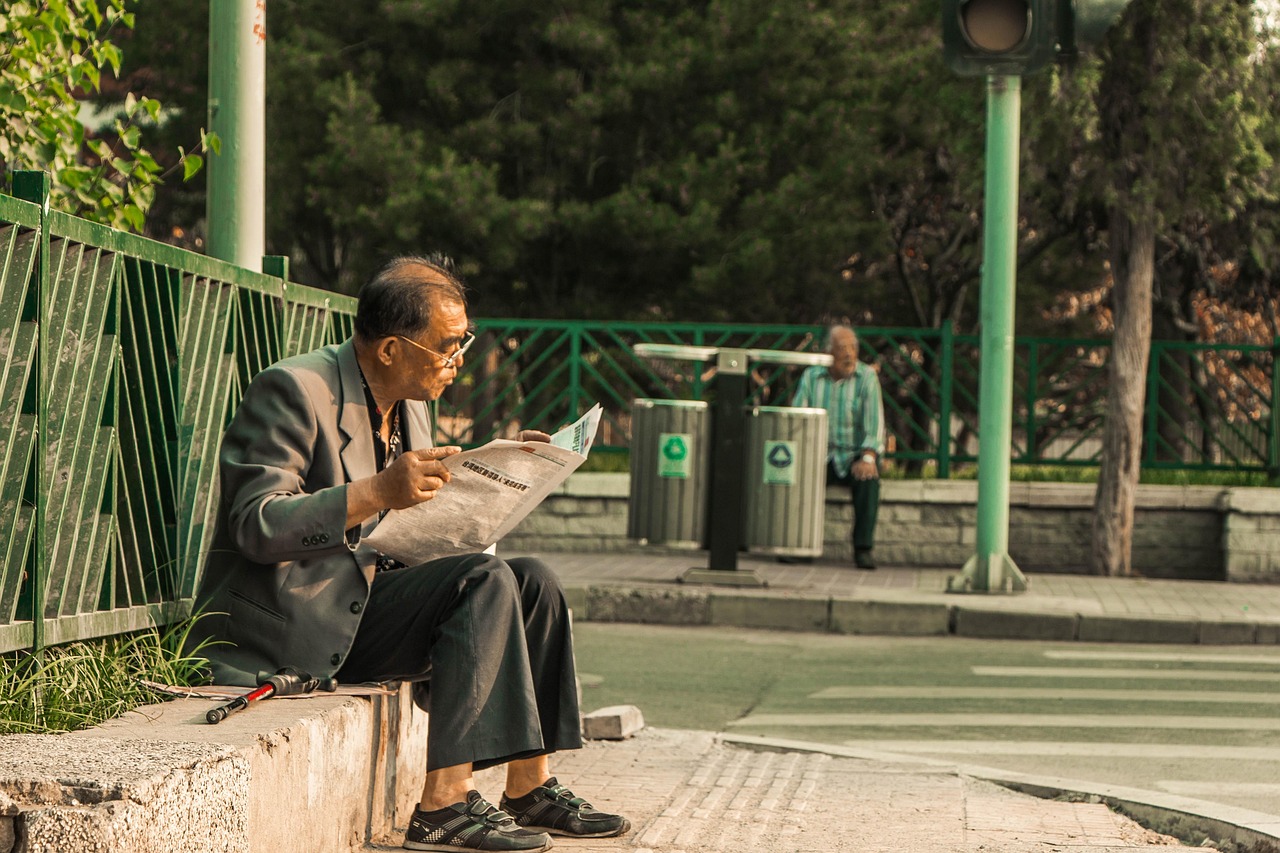 A man sits on a wall in the park and reads a newspaper. 