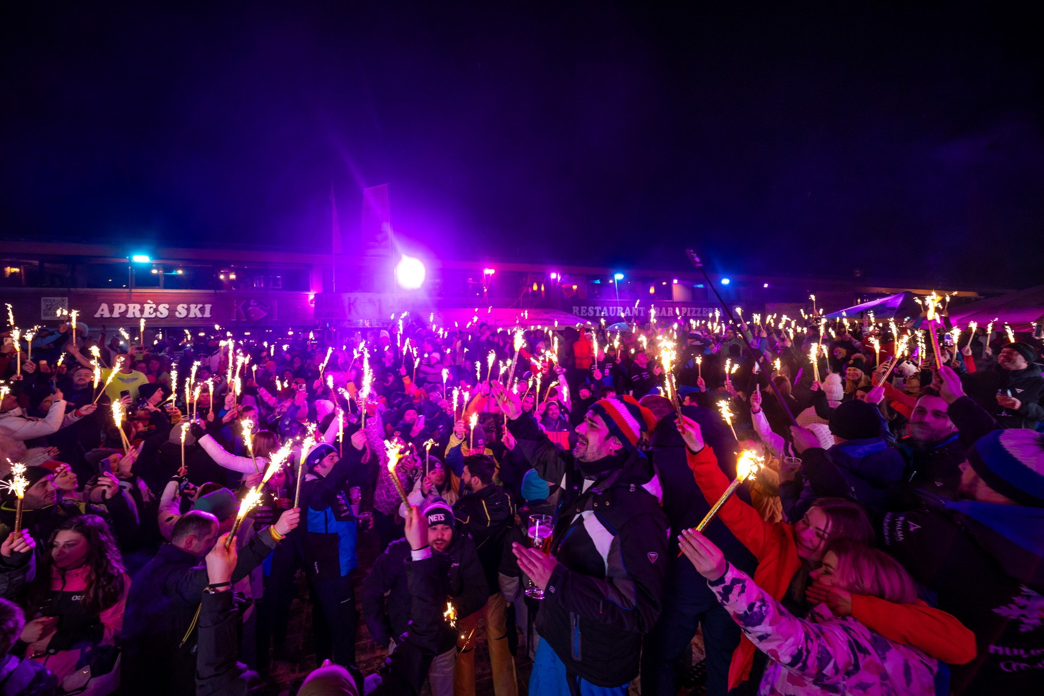 Large crowd enjoying a HULJS SKI apres-ski party at night with sparklers.